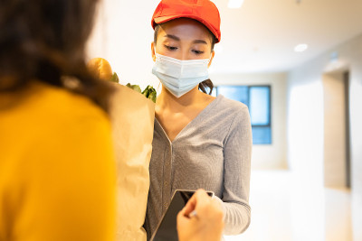 亚洲送人戴着口罩在红色制服处理袋食品，水果，蔬菜给公寓女顾客。新冠肺炎期间的邮差杂货送货服务。预防病