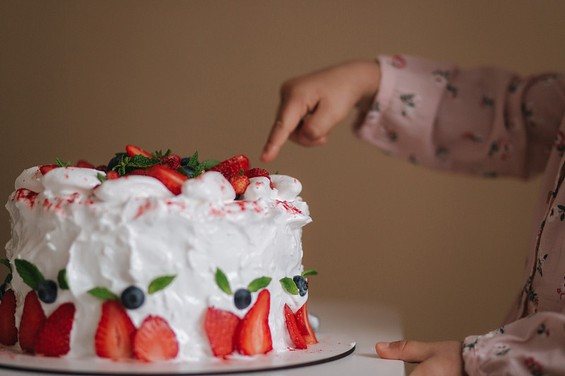 小女孩第一次品尝生日蛋糕。可爱的小女孩穿着漂亮的裙子品尝蛋糕和草莓。女孩的特写把手指放进蛋糕里