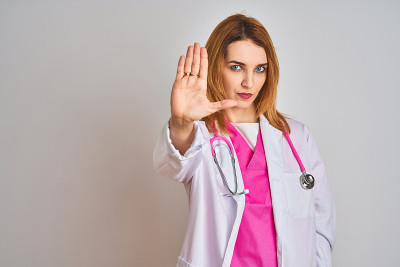 红发白种人医生女人穿粉红色听诊器在孤立的背景做停止唱歌与手掌。警告表情与否定和严肃的手势在脸上。