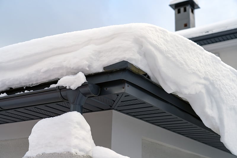 金属落水管系统，排水沟系统，雪下外部落水管和排水管。屋角的屋顶是灰色的金属瓦，排水沟在冬天覆盖着厚厚