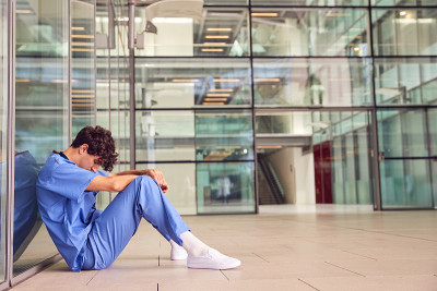 疲惫的年轻男医生穿着手术服坐在现代医院大楼靠墙