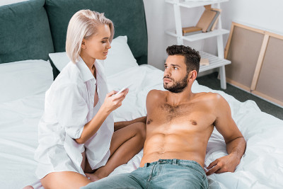 迷人的年轻女子坐在床上，用智能手机压着她的男友