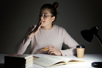迷人的学生闭着眼睛打哈欠，坐在桌面准备上课，加班。疲惫的女性坐在台灯旁，在黑暗的背景下喝着咖啡。
