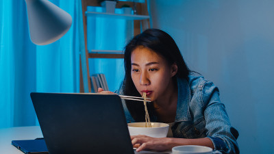 亚洲聪明的自由职业商业女性晚上在家里的客厅里用笔记本电脑工作的时候吃着方便面。