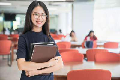 回到学校教育知识学院的大学理念，美丽的女大学生抱着她的书微笑着站在图书馆，学习和教育的理念