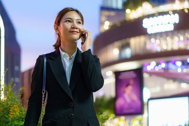 在泰国曼谷，一名亚洲行政工作女性晚上在以写字楼为背景的街道上使用手机。