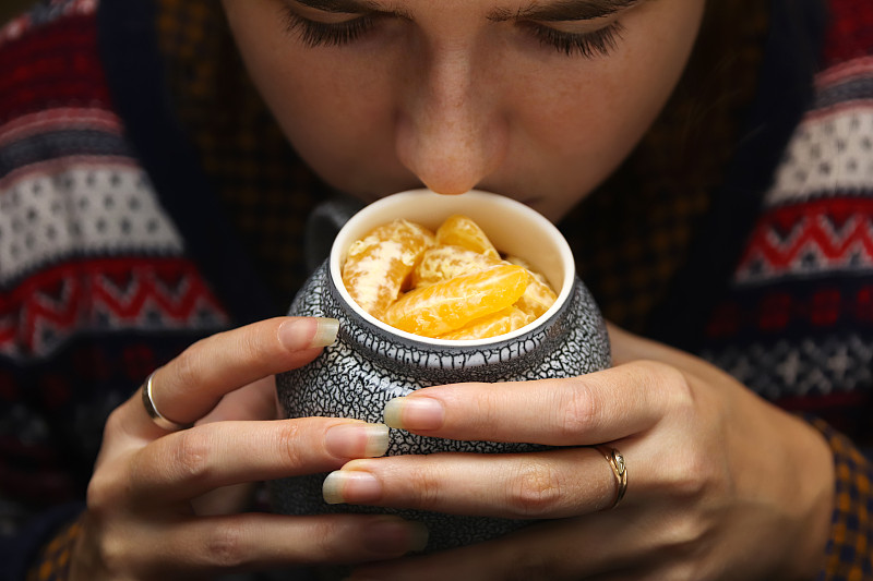 特写的女性手与一杯创意饮料桔子小叶。年轻的潮女穿着圣诞毛衣闻着水果的味道。舒适温暖的陶瓷。长指甲。冬