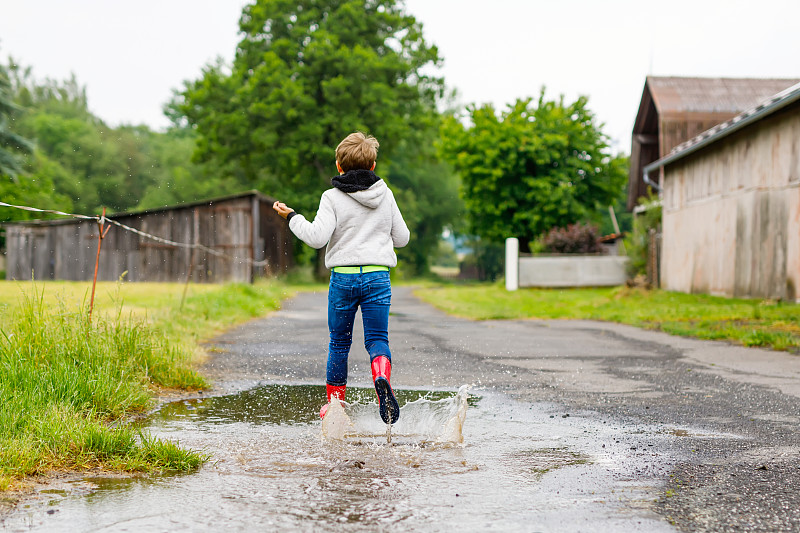 快乐的小男孩穿着红色雨靴，在雨夹雪和阴雨天行走。孩子在五颜六色的休闲衣服跳进水坑。在户外玩，健康的儿