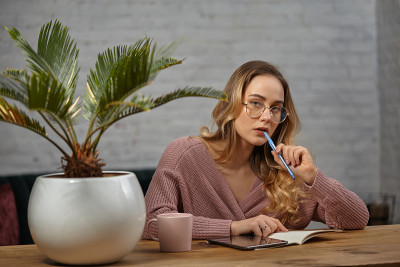 女孩博主穿着粉色开衫，戴着眼镜。坐在木桌旁，准备在笔记本上写点什么。花盆、杯子和牌匾旁的棕榈树
