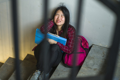 亚洲女大学生被欺负的戏剧性写照。年轻的沮丧和悲伤的日本女孩孤独地坐在校园楼梯遭受欺负和骚扰感到绝望和