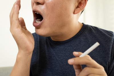 男人拿着一根香烟，用手检查他的呼吸是否有口臭，来自烟草烟雾的口腔异味，患牙周病的风险，口腔癌或吸烟引