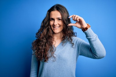年轻美丽的女人与卷发穿着蓝色休闲毛衣在孤立的背景微笑和自信的手势与手做小尺寸的手势与手指看和相机。测