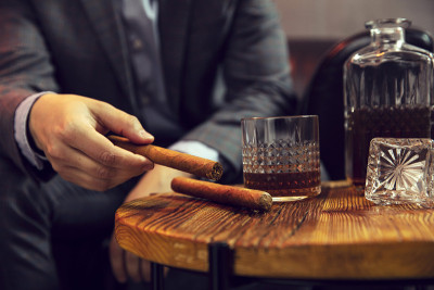 绅士从一张木桌上拿起一支雪茄，桌上放着一瓶威士忌和玻璃杯