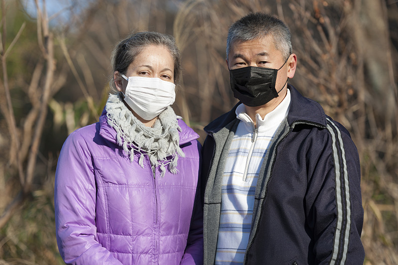 中年夫妇戴口罩(白色口罩和黑色口罩)，以预防病毒、流感、花粉热等疾病。看着摄像机。