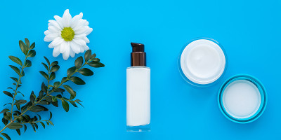 一套化妆品瓶子，面霜和乳液在一个蓝色的背景与一个绿色的树枝和雏菊。玻璃瓶内喷白洋甘菊，护肤理念。草本