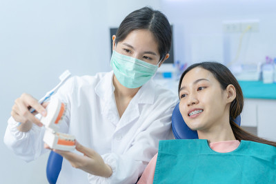 亚洲年轻女孩正在听女牙医讲解口腔护理和牙齿矫正患者的刷牙。牙医在牙科诊所佩戴防护口罩预防COVID-