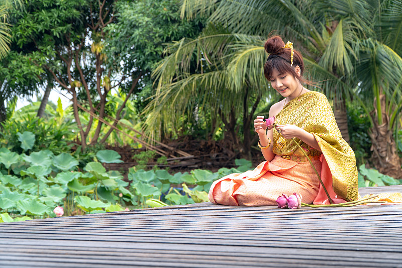 美丽的亚洲妇女穿着泰国传统服装，坐在木桥上。她的手是泰国风格的尊敬的手。侧面的荷叶上有一朵粉红色的荷