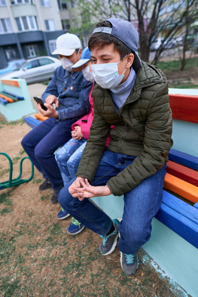 父亲和孩子们坐在有公寓的高层建筑附近操场上的长椅上，他们脸上戴着医用口罩，防止病毒和灰尘