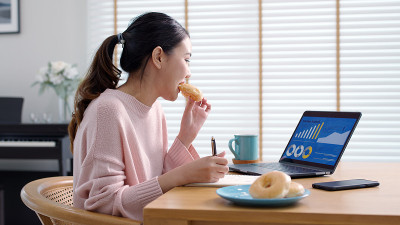 年轻迷人美丽的亚洲女性饿了吃甜甜圈，吃零食吃满嘴看电脑笔记本在家里在忙碌的工作，从家里多任务不健康的