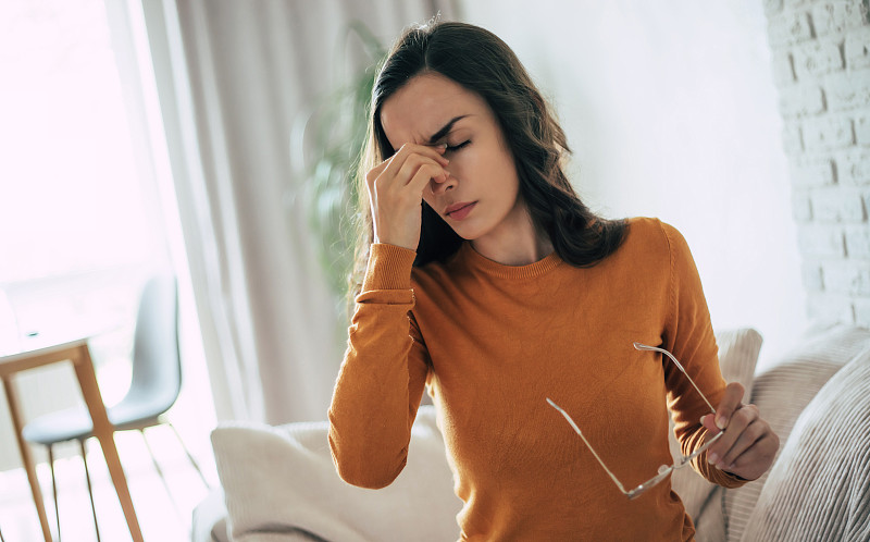 年轻悲伤的黑发女人患有严重的头痛，她坐在家里的沙发上