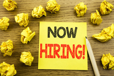 Word, writing Now Hiring。招聘的经营理念写在贴纸上的木制背景。背景是折叠的黄