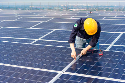 戴着黄色头盔的电工工程师正在检查建筑物屋顶上的太阳能电池板。
