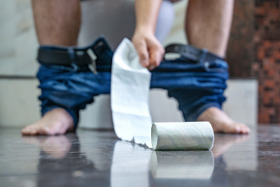 一个男人坐在浴室的马桶上，伸手去拿手纸。腹泻胃痛。