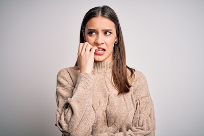 年轻美丽的黑发女人穿着休闲毛衣站在白色背景看起来紧张和紧张的手在嘴咬指甲。焦虑的问题。