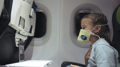 白人小女孩戴着防护面具在飞机上。婴儿游客在飞机上戴着呼吸器玩智能手机。新型冠状病毒肺炎