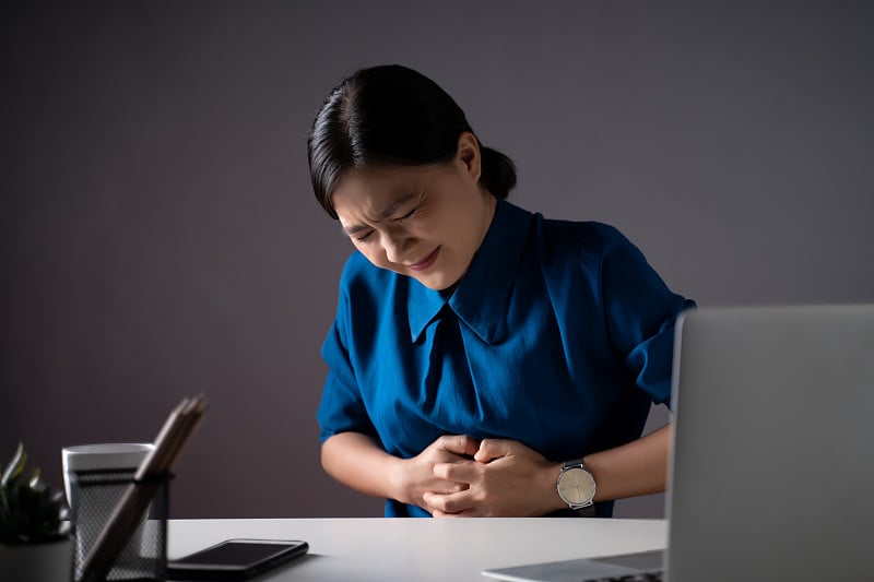 一名身穿蓝色衬衫的亚洲妇女坐在办公室里胃痛。孤立在白色背景上。