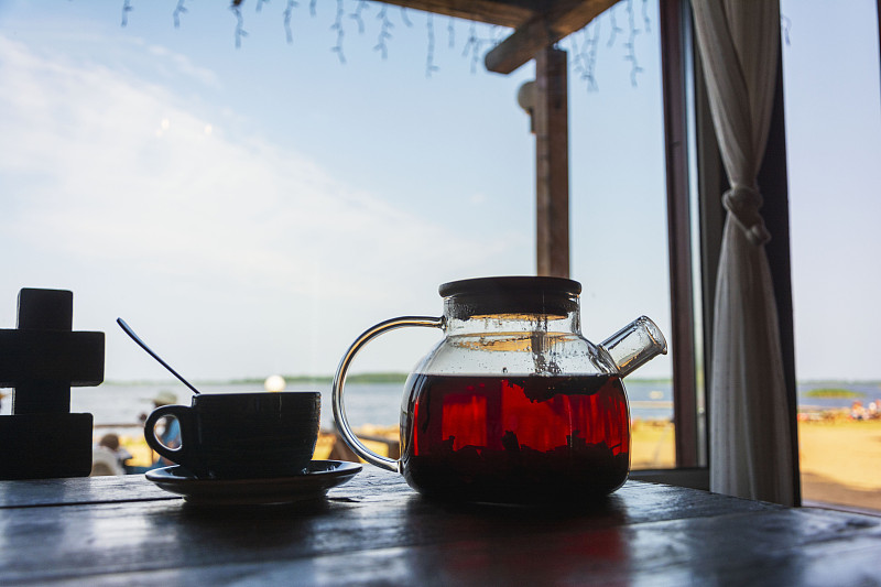 茶装在玻璃茶壶里，茶杯放在户外的木桌上。沿海咖啡馆,早午餐。海滩和大海都模糊了