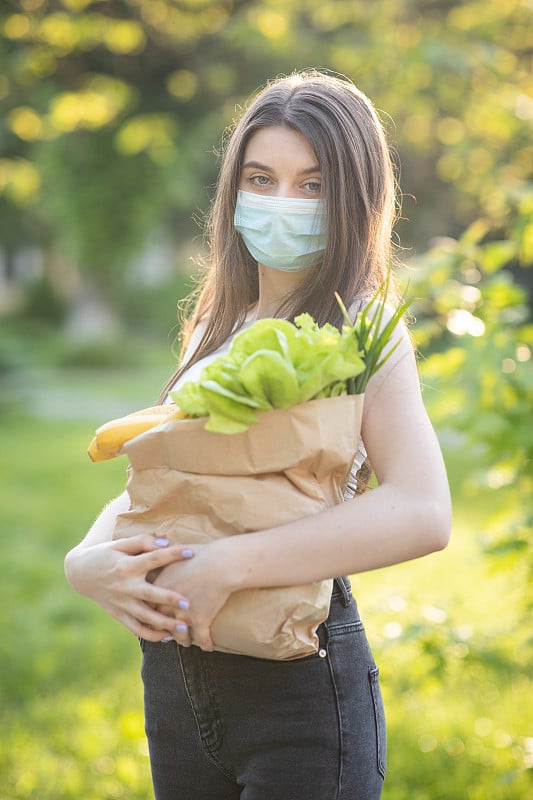 白种年轻漂亮的女送货员在公园里戴着医用口罩，带着一包新鲜食物的垂直肖像