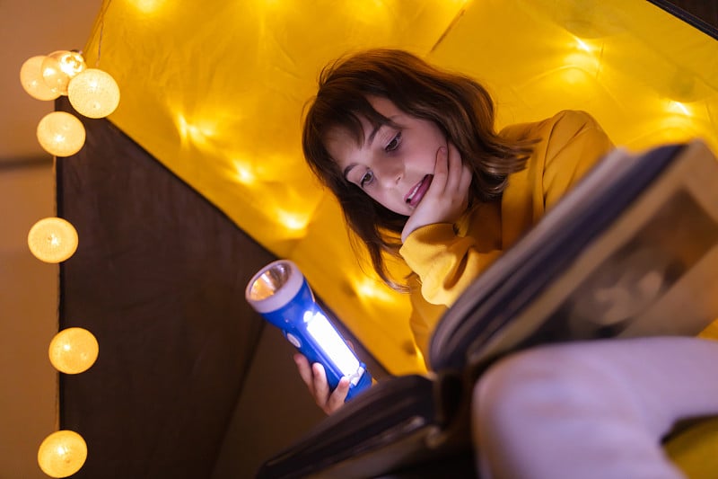 年轻的女孩子在家里用轻球做的起居室帐篷里阅读百科全书。