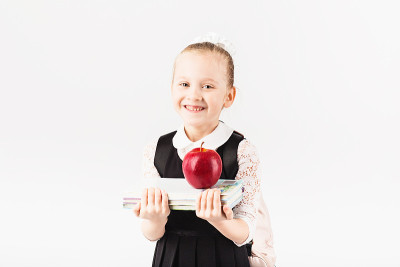 书、学校、孩子。小女孩面带微笑，背着大背包，白色背景下拿着书和大红苹果。