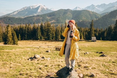 年轻的金发女子拿着保温杯在山里用智能手机聊天。自由，快乐，旅游和度假的概念，户外活动。快乐女孩戴着红