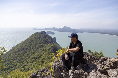 在泰国的普拉卓布基里汗省，亚洲男人坐在山顶上欣赏海景。穿着黑衣服的男人在山顶上看大海的景色。