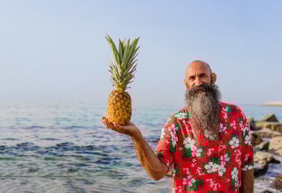一个留着漂亮胡子的秃顶老男人，穿着一件夏威夷衬衫，在海滩上拿着一个菠萝