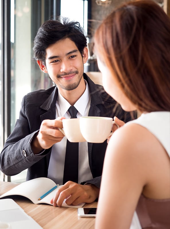 两个年轻的商人/女人在休闲西装聊天和喝咖啡在咖啡馆会面