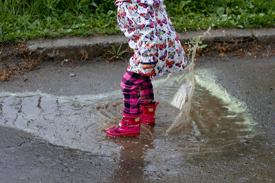 小女孩穿着五颜六色的雨衣和雨靴在泥泞的水坑里跳