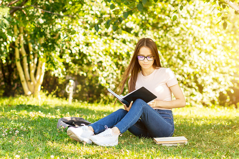 少年坐在草地上学习，一个女学生在大学公园的树下背着书包和课本