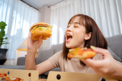 饥饿的年轻女人吃垃圾食品汉堡和披萨午餐订购送货上门度假。不健康饮食，肥胖风险。