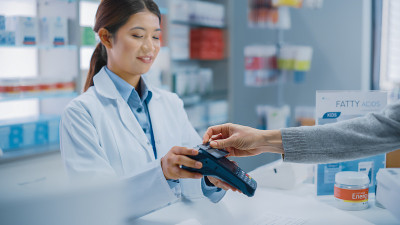 药店收银台:亚洲职业女性药剂师销售打包药品，白人女性客户使用非接触式支付终端和信用卡支付