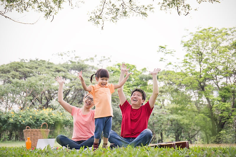 幸福家庭的肖像男人女人和小女孩在外面玩。年轻的三口之家一起在户外野餐。亚洲的父母和女孩高兴地跳和微笑