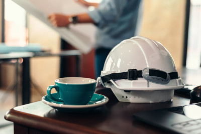 安全帽、咖啡杯放在工地施工桌上。建筑师、工程师读规划模糊背景。