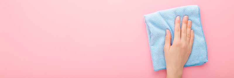 年轻的成年妇女手拿蓝色抹布，擦拭厨房、浴室或其他房间的桌子、墙壁或地板表面。亮粉红色背景。柔和的颜色