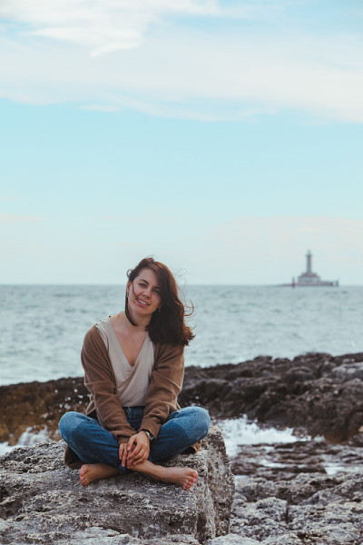一个女人坐在布满岩石的海滩上，穿着湿牛仔裤，背景是灯塔