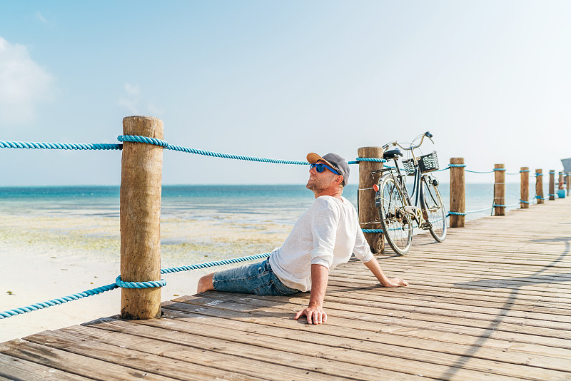 肖像放松的男人穿着轻夏装和太阳眼镜坐和享受时间和海滩上的木制码头。热带国家无心度假的概念形象。坦桑尼