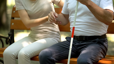 女性帮助盲人拄着拐杖坐在长椅上，志愿者服务近距离