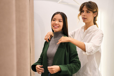 年轻的时尚顾问自信地帮助漂亮的亚洲女孩在现代商店试穿开襟羊毛衫
