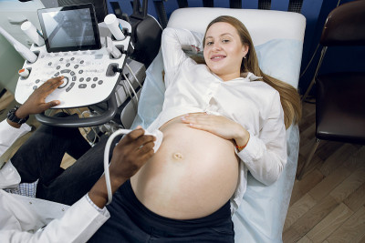 俯视图微笑愉快的怀孕金发白种人女人可爱的肚子，在现代妇科和产科诊所有超声波扫描。男非洲医生的手做超声